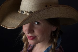 Head and shoulders photographic portrait of blonde model in plaits wearing cowboy hat looking cheeky